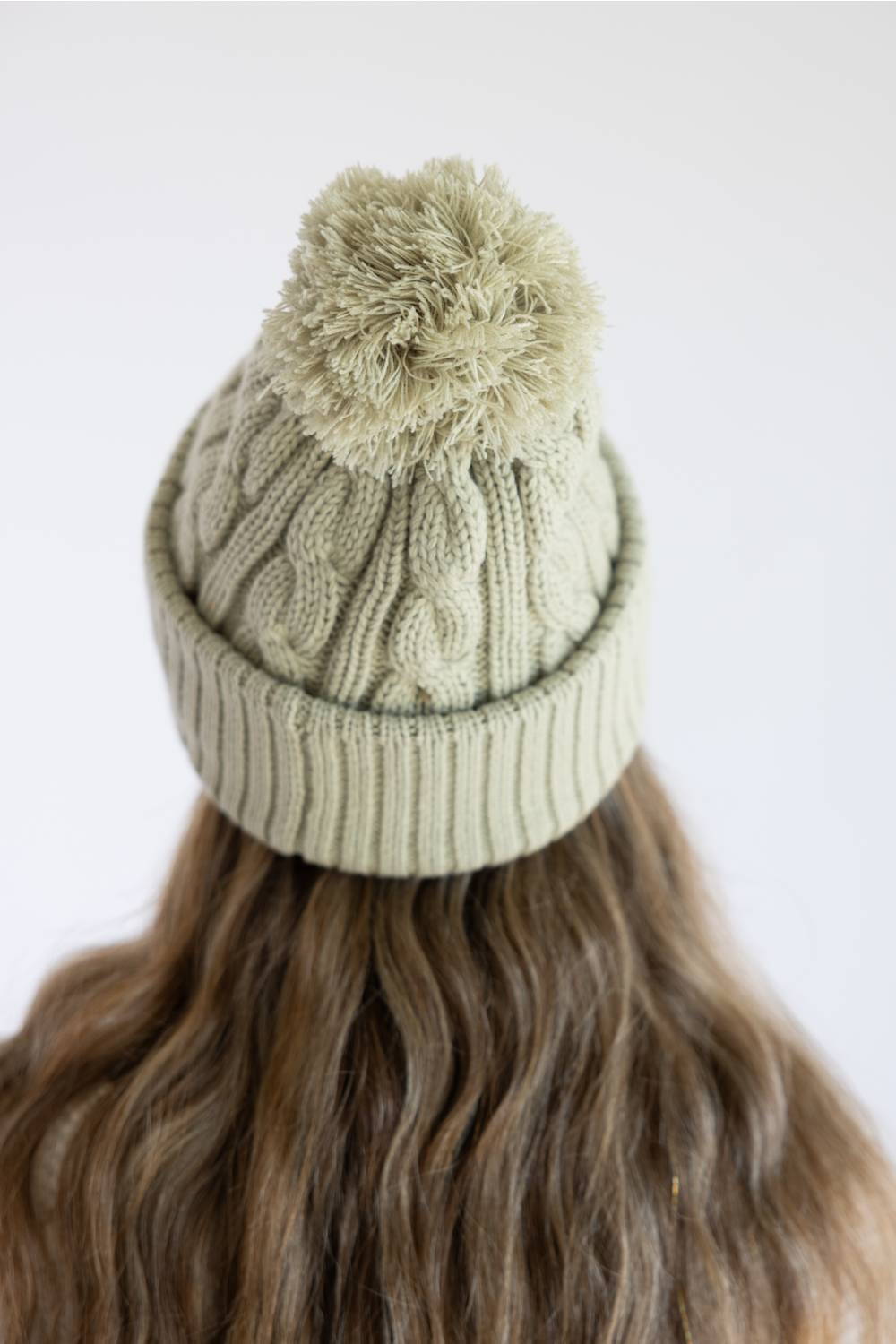 A person viewed from the back wearing a light sage green cable-knit beanie with a matching pom-pom, against a plain white background.