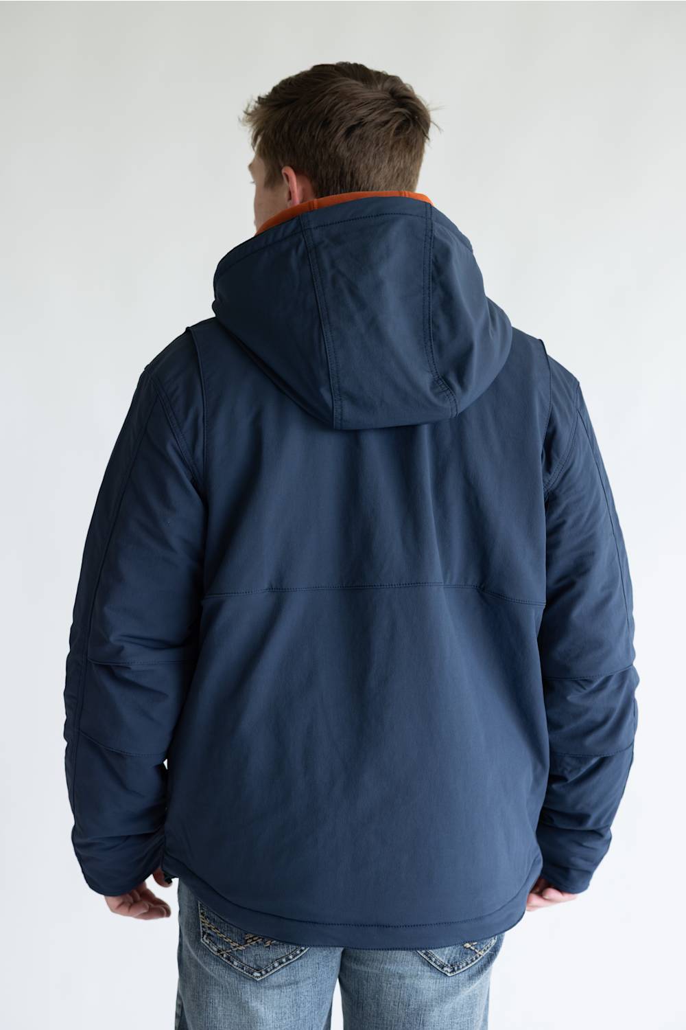 A man viewed from behind wearing a navy blue hooded jacket with a brown collar lining and blue jeans, set against a plain light gray background.