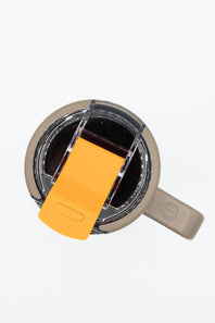 Top-down view of a beige insulated tumbler with a clear lid and a bright orange flip-up straw cover, shown on a white background.