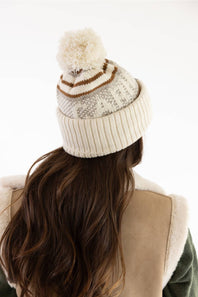 Rear view of a model with long brunette hair wearing a Carhartt thick knit cuffed beanie with a pom-pom in Oat Milk and a beige sherpa-lined vest