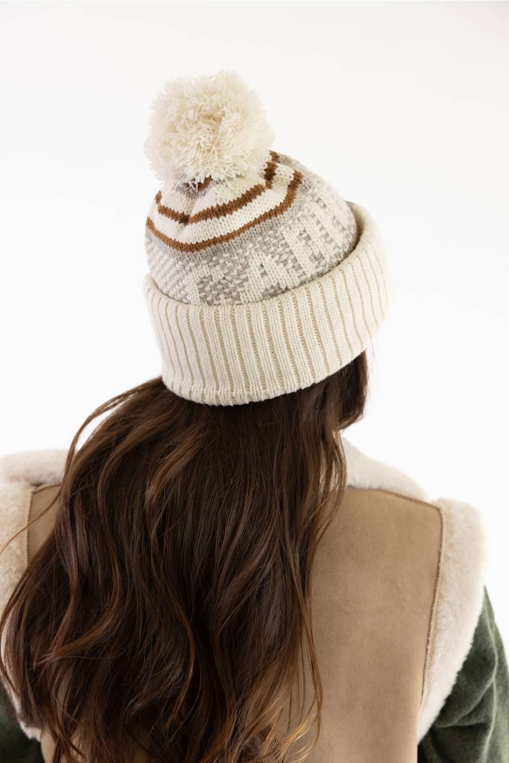 Rear view of a model with long brunette hair wearing a Carhartt thick knit cuffed beanie with a pom-pom in Oat Milk and a beige sherpa-lined vest