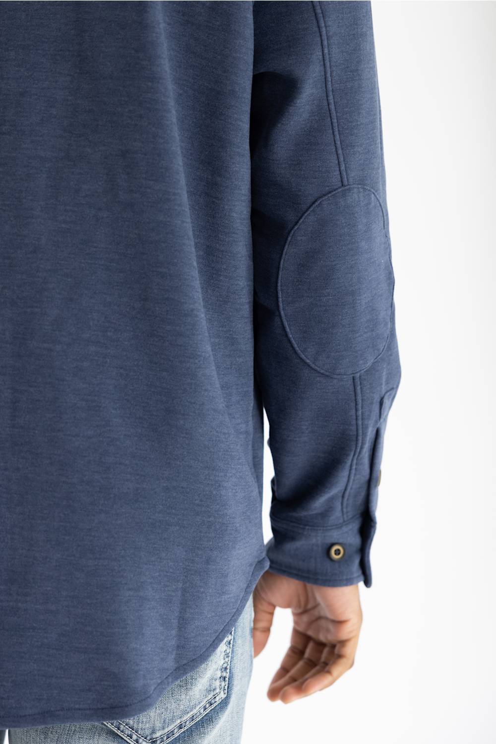 Close-up of a person wearing a heathered blue long-sleeve shirt with a round elbow patch and a buttoned cuff, paired with light blue jeans against a white background.