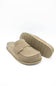 A pair of taupe suede mule slip-on shoes, with one shoe standing upright to show the sole and the other lying flat to show the top, set against a plain white background.