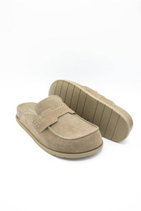 A pair of taupe suede mule slip-on shoes, with one shoe standing upright to show the sole and the other lying flat to show the top, set against a plain white background.