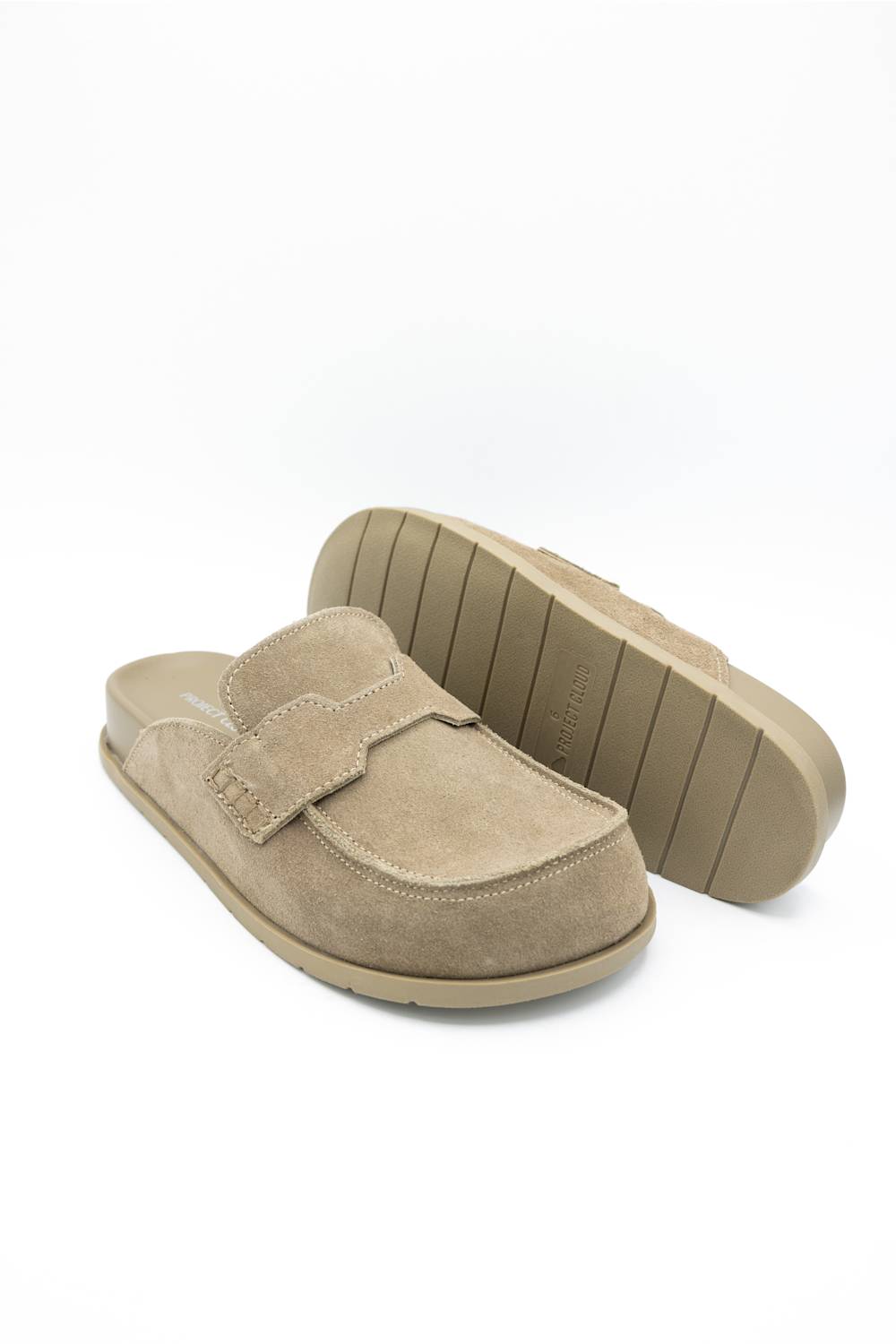 A pair of taupe suede mule slip-on shoes, with one shoe standing upright to show the sole and the other lying flat to show the top, set against a plain white background.