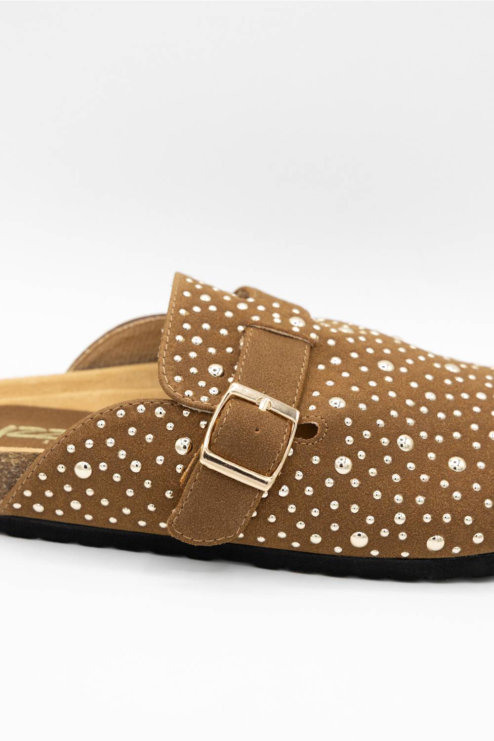 A close-up of a brown suede clog-style mule sandal decorated with small white rhinestones, featuring a wide strap with a gold-tone buckle.