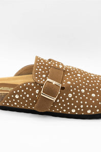 A close-up of a brown suede clog-style mule sandal decorated with small white rhinestones, featuring a wide strap with a gold-tone buckle.