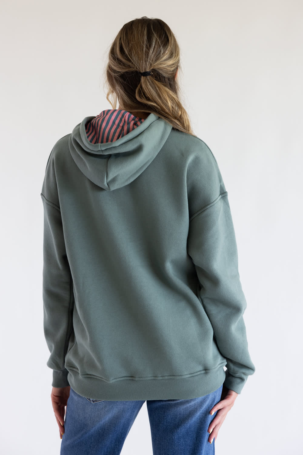 A woman viewed from the back wearing an oversized sage green grey   hoodie with a striped lining inside the hood, paired with blue jeans, against a plain white background.