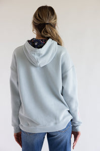 A woman viewed from behind wearing a light blue hooded sweatshirt with a floral-patterned inner hood lining and blue jeans, set against a plain off-white background.