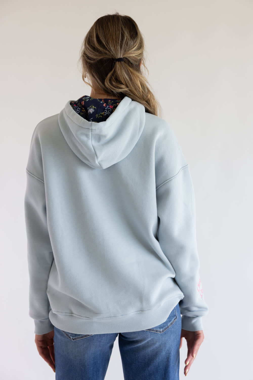 A woman viewed from behind wearing a light blue hooded sweatshirt with a floral-patterned inner hood lining and blue jeans, set against a plain off-white background.
