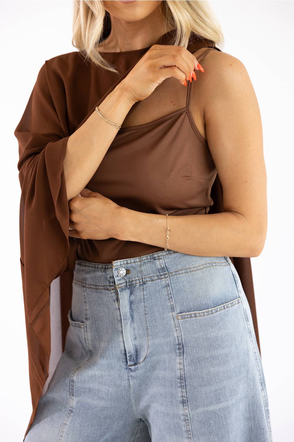 A woman wearing a brown sleeveless camisole top with sheer mesh panels and a draped brown shawl, paired with light blue jeans, against a plain white background.