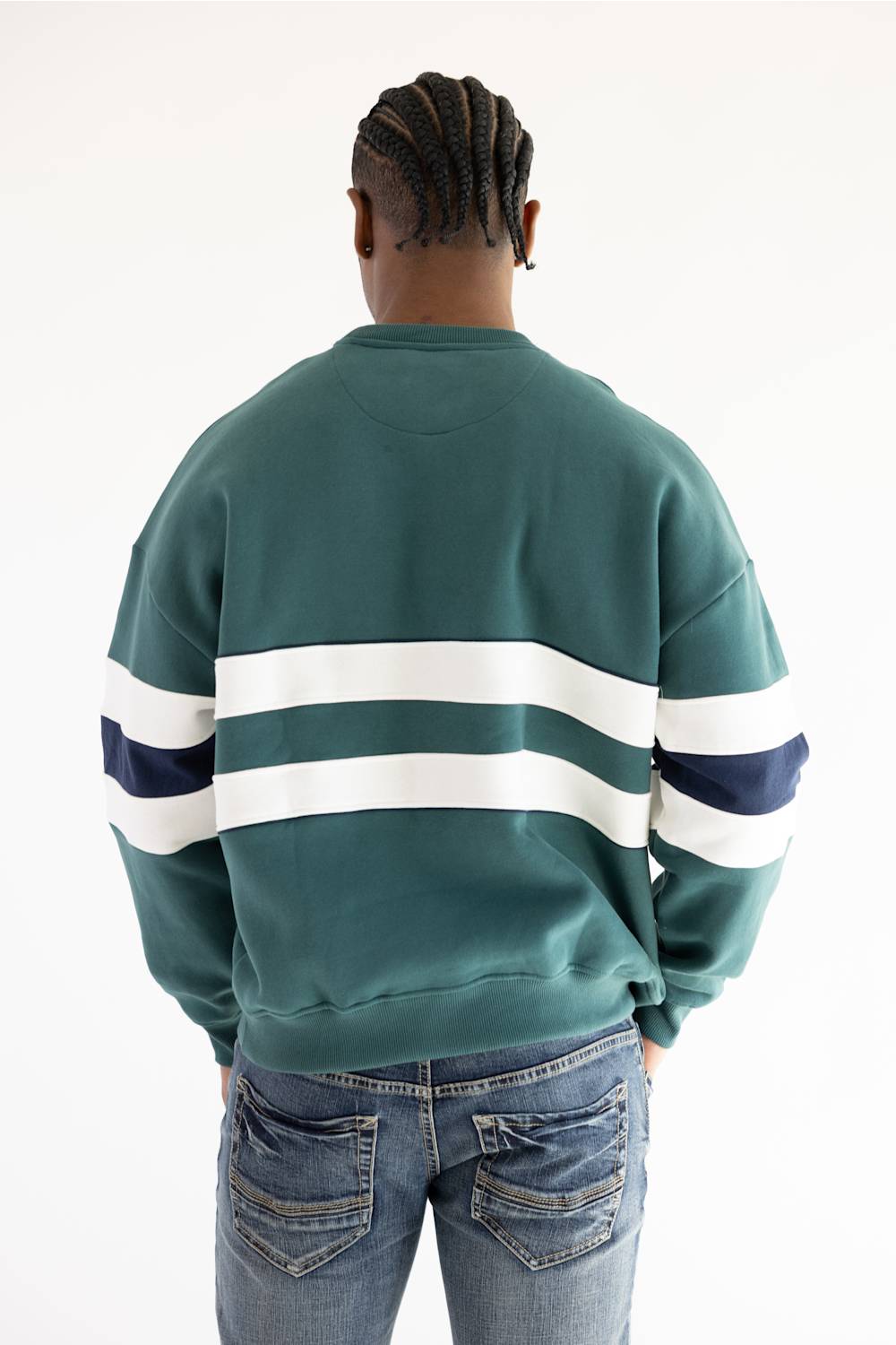A person viewed from the back wearing a green crewneck sweatshirt with two horizontal white stripes across the shoulders and upper arms, plus a dark navy stripe at the end of each white stripe. The person is also wearing blue jeans against a plain white studio background.
