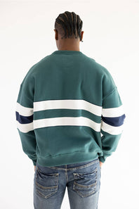 A person viewed from the back wearing a green crewneck sweatshirt with two horizontal white stripes across the shoulders and upper arms, plus a dark navy stripe at the end of each white stripe. The person is also wearing blue jeans against a plain white studio background.