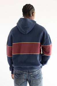 A person viewed from the back wearing a navy hoodie with a wide burgundy horizontal stripe across the upper back and sleeves, outlined with thin gold trim, paired with blue jeans against a plain white background.