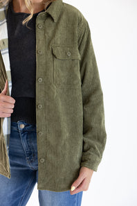 An olive green corduroy button-up shirt jacket worn by a model, styled over a black top and blue jeans against a plain white background.