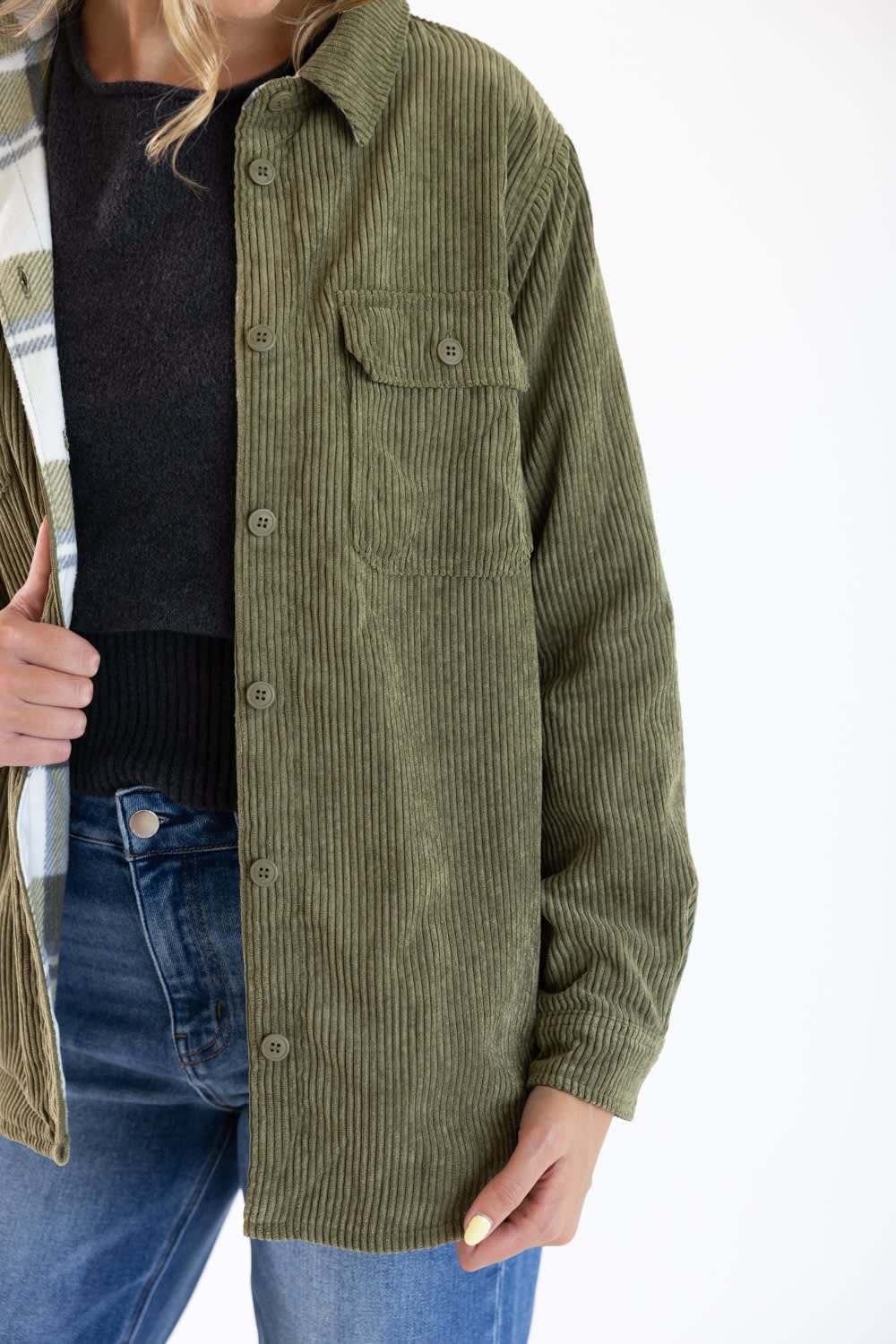 An olive green corduroy button-up shirt jacket worn by a model, styled over a black top and blue jeans against a plain white background.