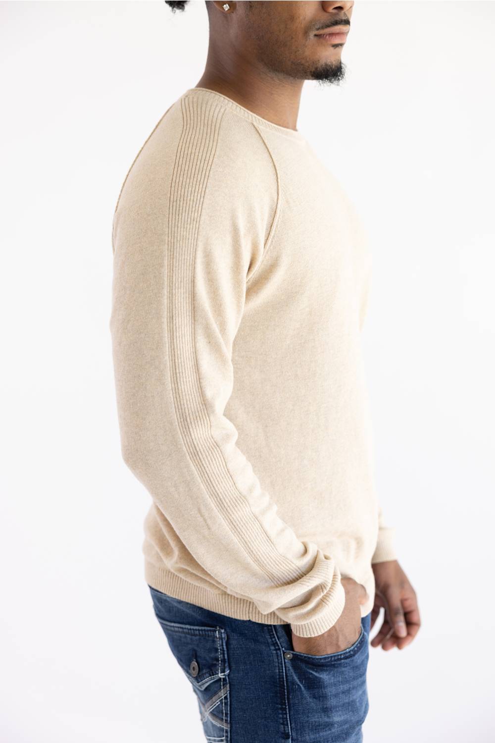 A man shown in profile wearing a light beige ribbed knit sweater and blue jeans against a plain white studio background.