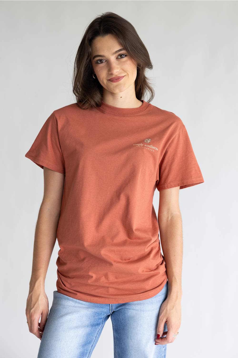 A woman wearing a rust-colored short-sleeve t-shirt with a small logo on the left chest, paired with light blue jeans, standing against a light gray background.