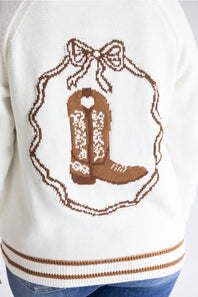 Close-up of a white knit sweater with a brown embroidered cowboy boot design inside a bow-shaped frame, with ribbed brown and white cuffs and blue jeans visible at the bottom.