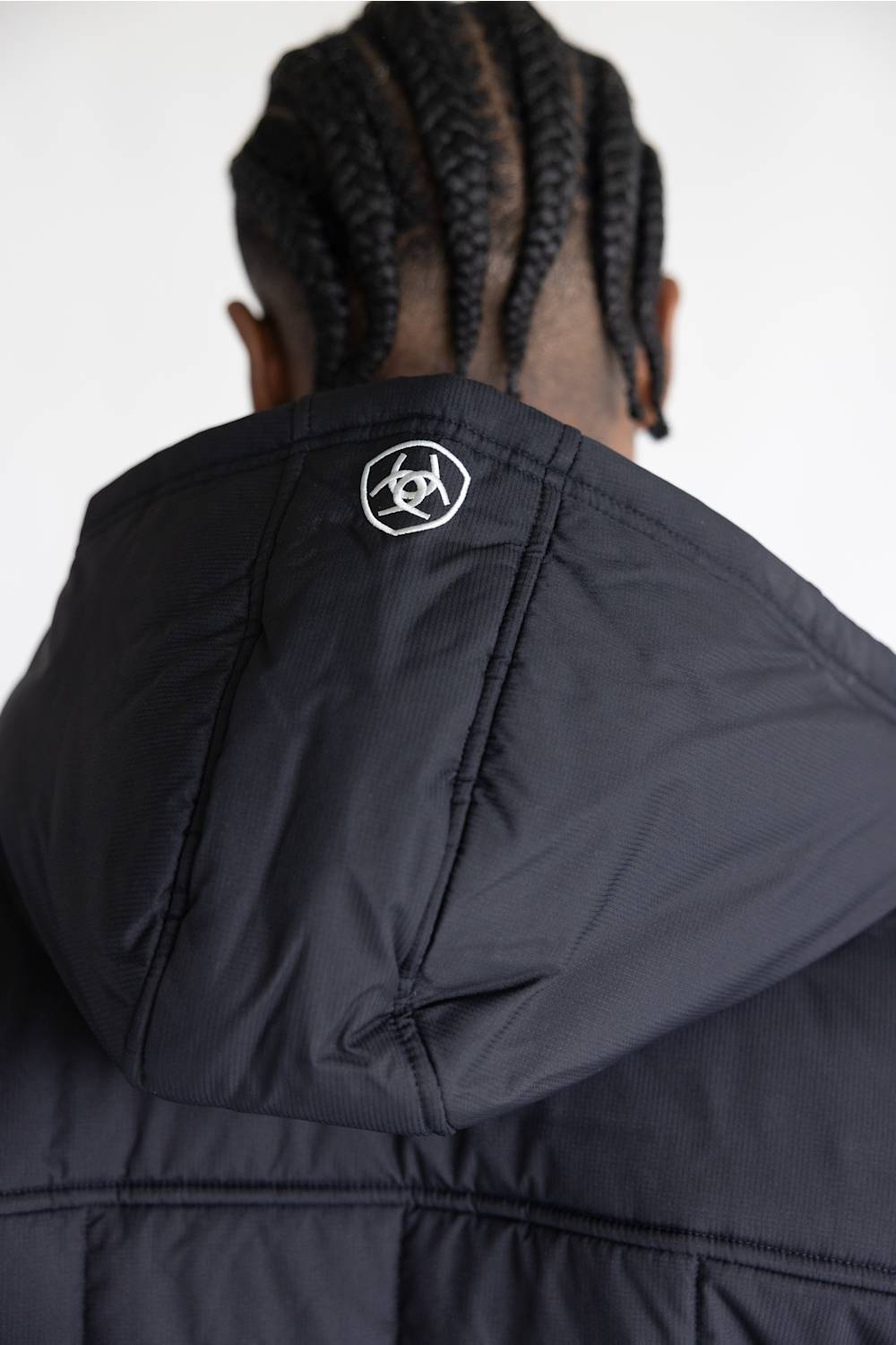 Close-up back view of a person wearing a black puffer jacket with a hood, showing a small white geometric logo on the back of the neck. The background is a clean white studio backdrop.