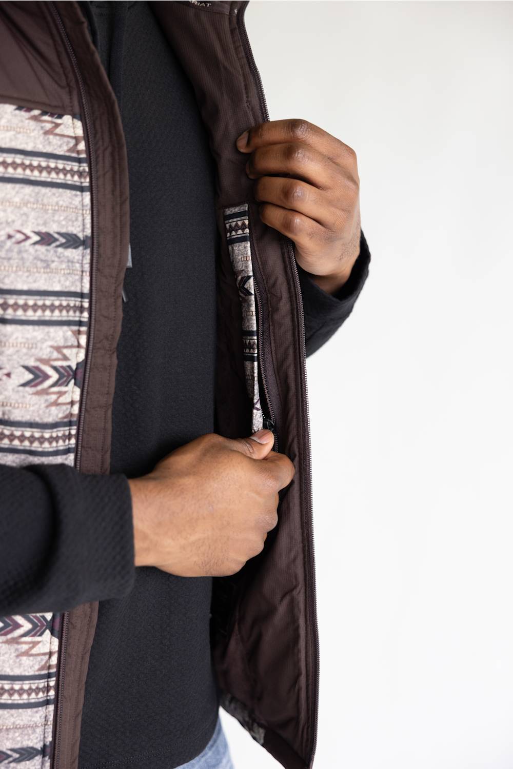 Close-up of a person wearing a dark brown puffer vest over a black sweater and patterned shirt, showing the zipper and inner lining.