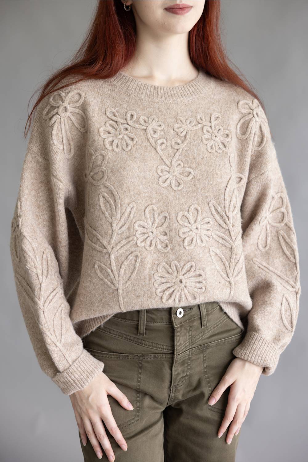 A woman wearing a cropped, light beige knit sweater with raised floral patterns and olive green pants, shown from the mouth down against a plain gray studio background.