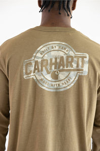 A close up of a man viewed from the back wearing a long sleeve light brown t-shirt with a graphic that says "CARHARTT BUILD BY HAND MADE WITH HEART" with a Carhartt logo C.