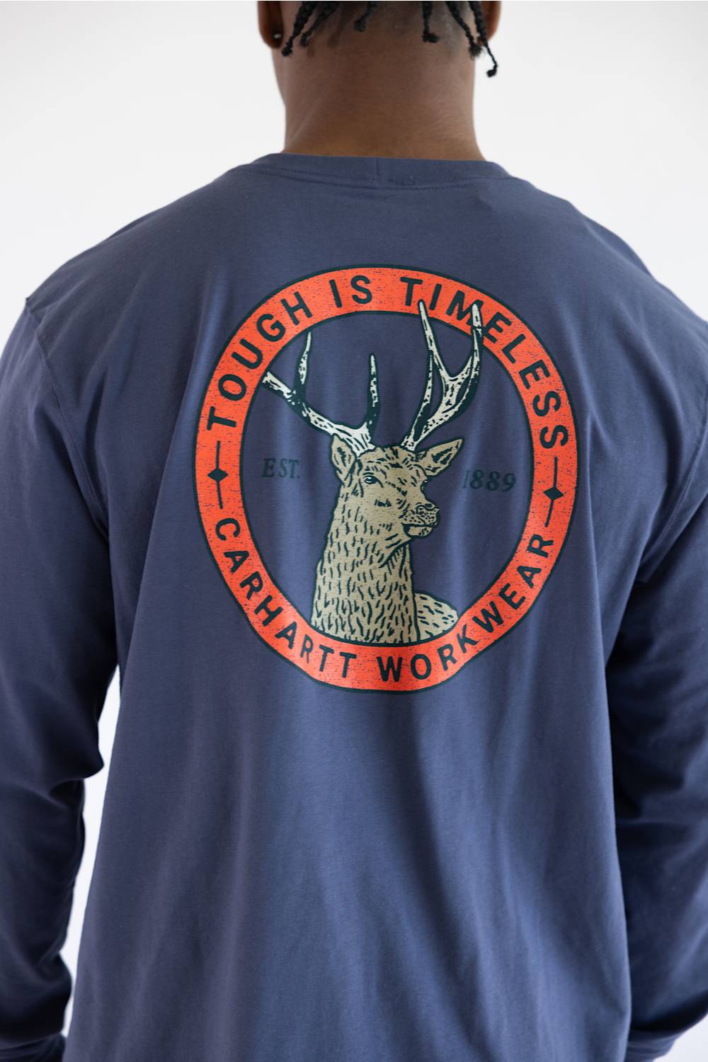 A close up of a man viewed from behind wearing a navy blue long-sleeve t-shirt with a large circular back graphic featuring a deer head and the text 'TOUGH IS TIMELESS' and 'CARHARTT WORKWEAR'.