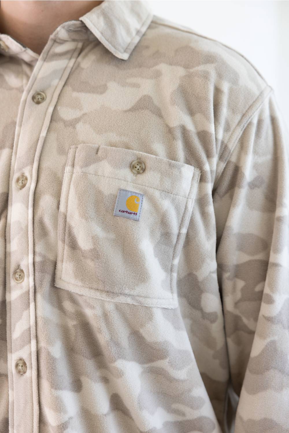 Close-up of a person wearing a Carhartt button-up shirt in a light beige and gray camouflage pattern, featuring a chest pocket with a visible Carhartt logo patch.