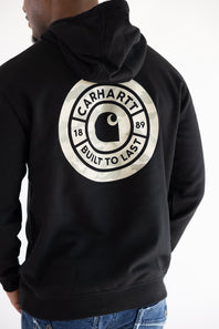 Detailed: Back view of a man wearing a black Carhartt Sawyer hoodie featuring a circular camo back graphic with the Carhartt logo and "1889 Built To Last" text.