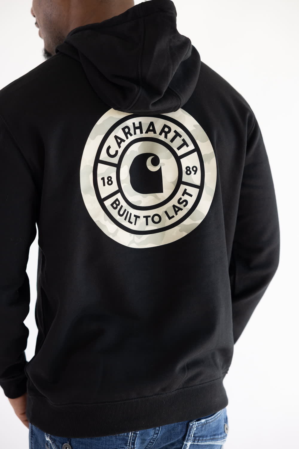 Detailed: Back view of a man wearing a black Carhartt Sawyer hoodie featuring a circular camo back graphic with the Carhartt logo and "1889 Built To Last" text.