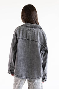 A person shown from the back wearing an oversized washed gray denim jacket, paired with light gray patterned pants, against a plain white background.