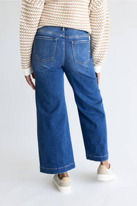A person shown from the back wearing blue wide-leg cropped jeans, a cream and brown knit sweater, and beige sneakers against a plain light gray studio background.