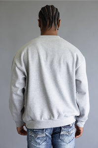 A man viewed from behind wearing a light gray crewneck sweatshirt and blue jeans with decorative stitching on the back pockets, set against a plain gray background.