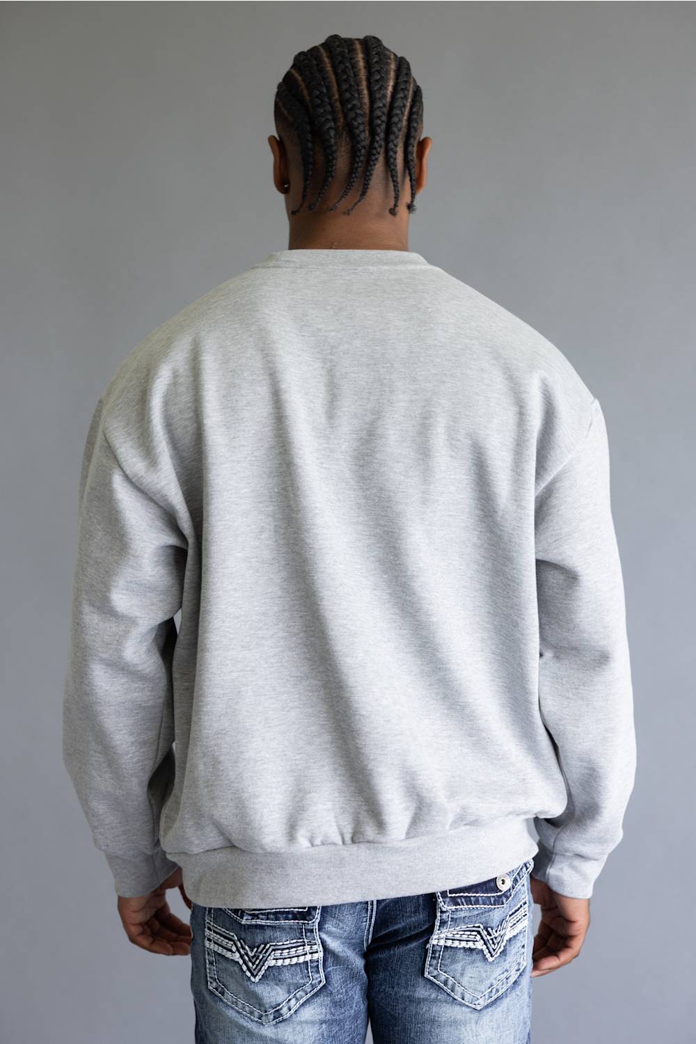 A man viewed from behind wearing a light gray crewneck sweatshirt and blue jeans with decorative stitching on the back pockets, set against a plain gray background.