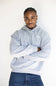 A man wearing a light gray heathered hoodie with a small white embroidered logo on the left chest, paired with blue jeans, standing against a plain white background.