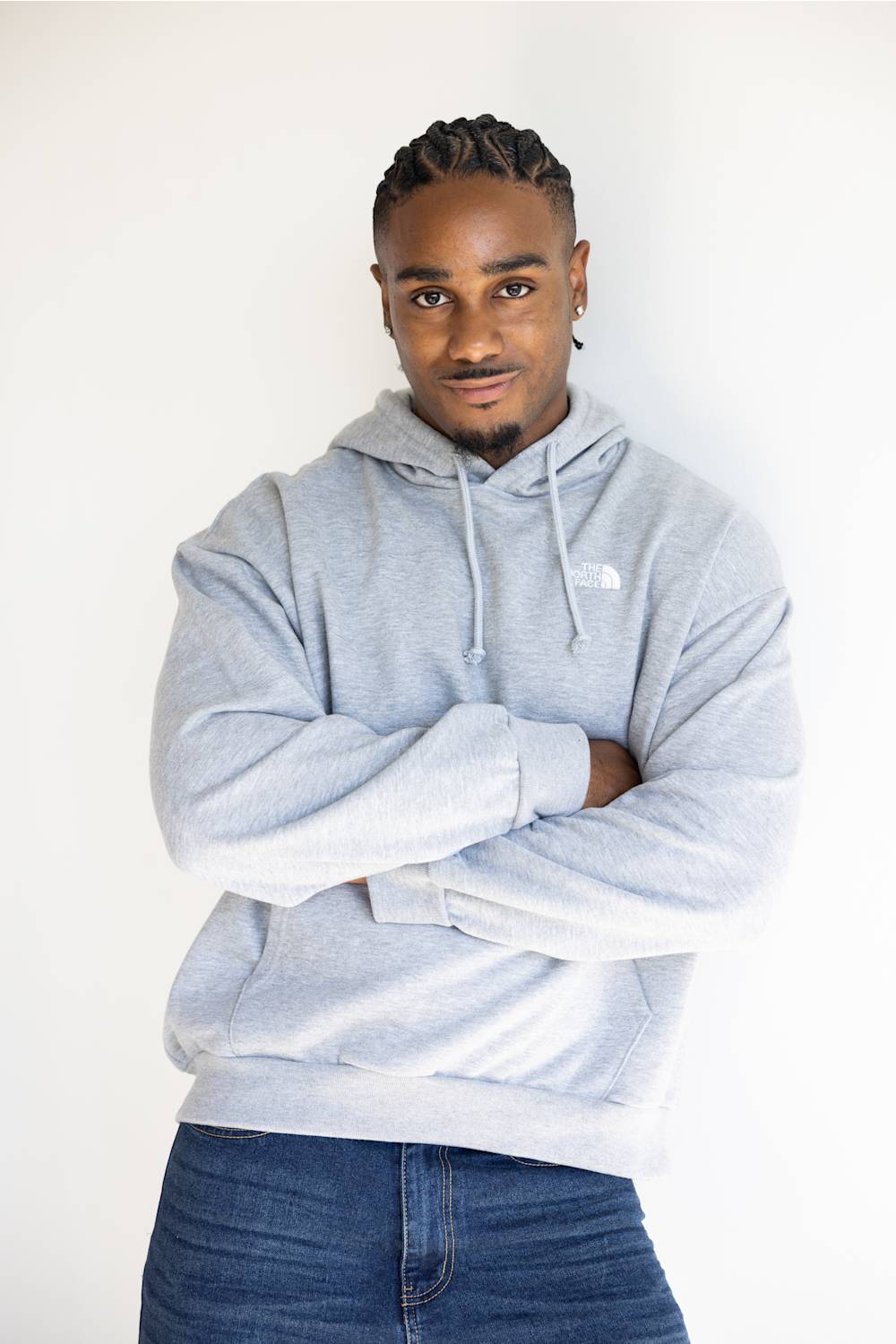 A man wearing a light gray heathered hoodie with a small white embroidered logo on the left chest, paired with blue jeans, standing against a plain white background.