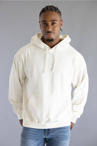 A man wearing a cream-colored pullover hoodie with drawstrings and a front kangaroo pocket, paired with blue jeans, standing against a solid gray studio background.