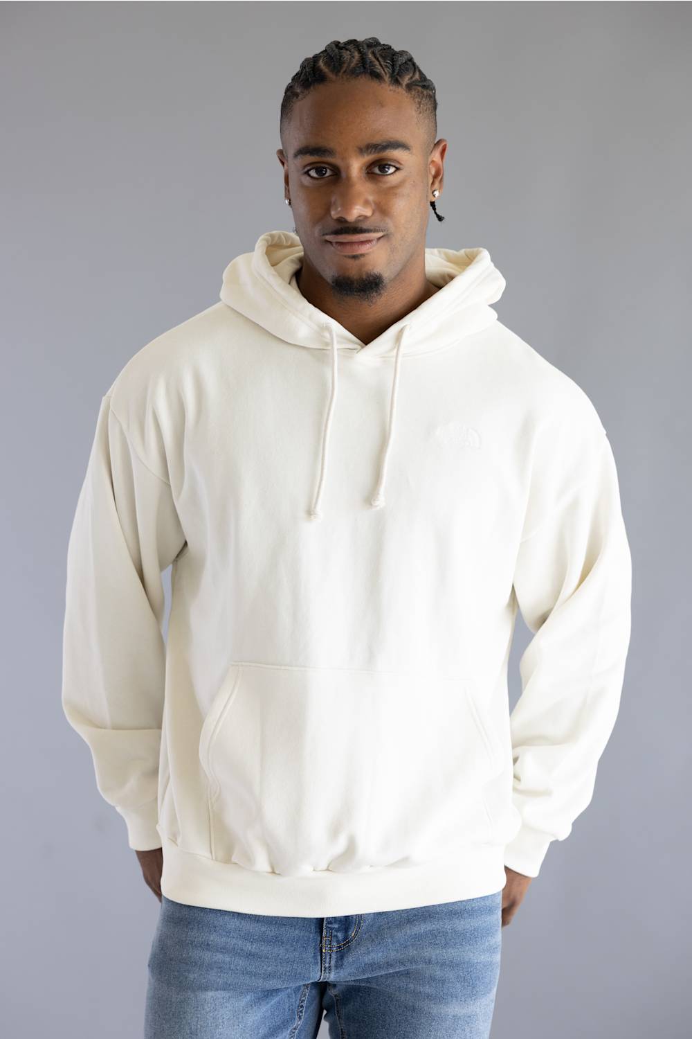 A man wearing a cream-colored pullover hoodie with drawstrings and a front kangaroo pocket, paired with blue jeans, standing against a solid gray studio background.