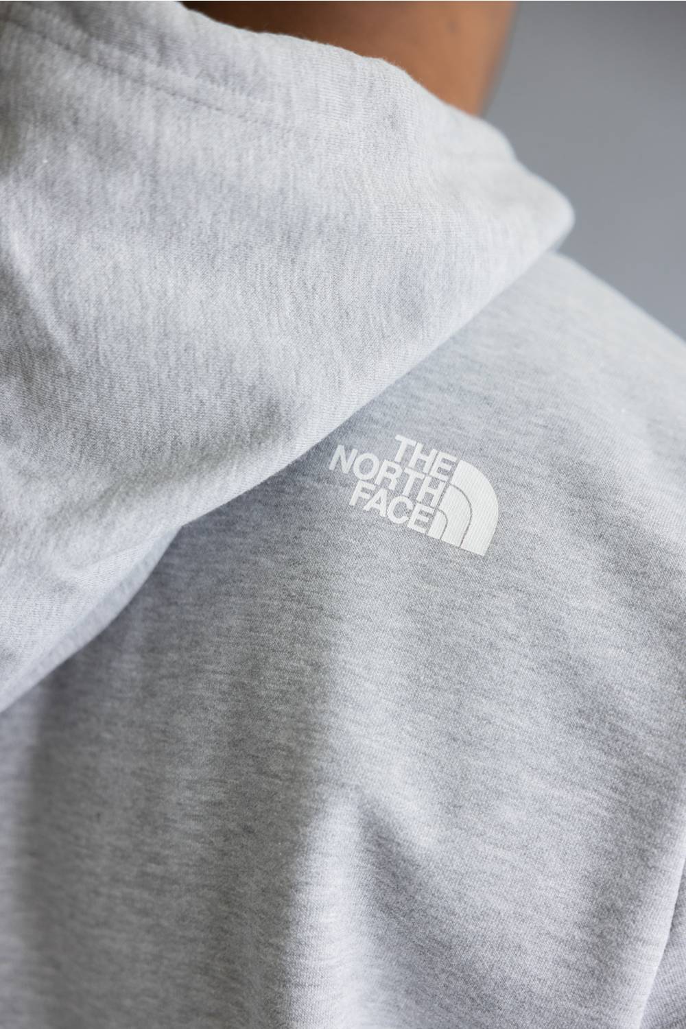 A man wearing a light gray heather pullover hoodie with a red 'The North Face' logo patch on the left chest, paired with blue jeans, against a solid gray studio background.