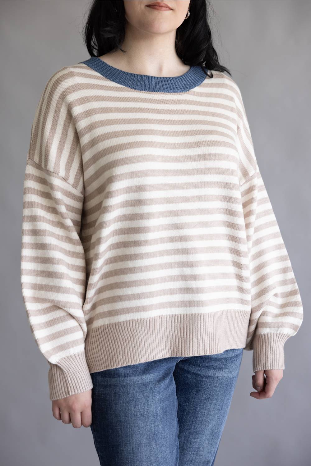 A woman wearing a beige and white horizontally striped knit sweater with a contrasting blue crew neck, paired with blue jeans, against a plain gray background.