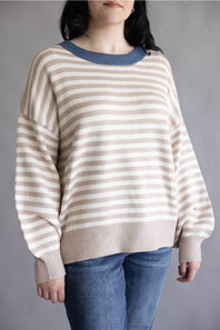 A woman wearing a beige and white horizontally striped knit sweater with a contrasting blue crew neck, paired with blue jeans, against a plain gray background.