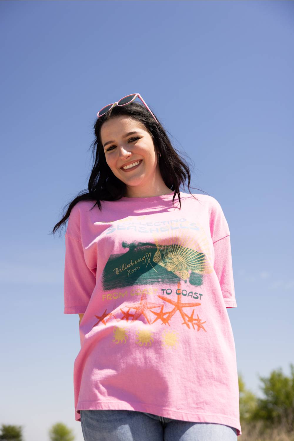 A woman wearing a pink Billabong graphic t-shirt with a green turtle and orange starfish, photographed outdoors against a clear blue sky.