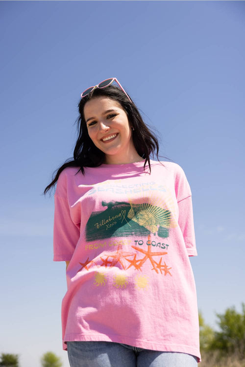 A woman wearing a pink Billabong graphic t-shirt with a green turtle and orange starfish, photographed outdoors against a clear blue sky.