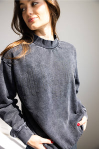 A woman wearing a dark gray, washed cotton crewneck sweatshirt with tonal embroidered lettering on the front, posed against a neutral gray background.