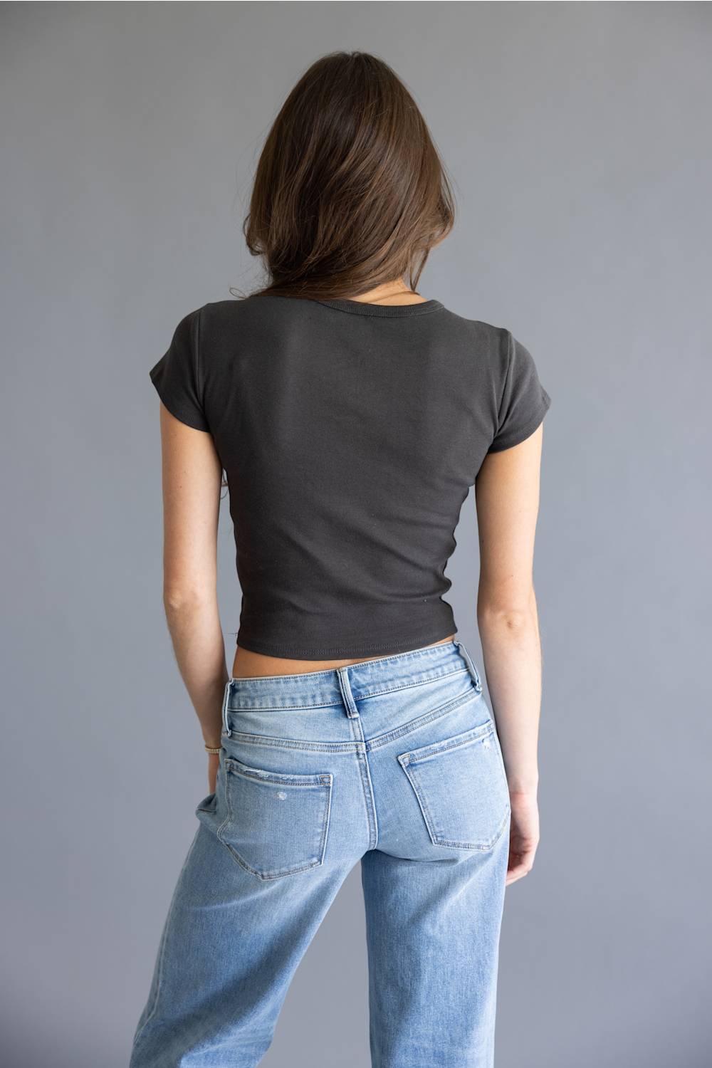 A woman shown from behind wearing a dark charcoal gray short-sleeve crop top and light blue jeans, standing against a solid gray studio background.