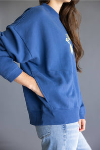 A person wearing an oversized blue sweatshirt with a small white graphic on the chest, paired with light blue jeans, shown against a plain gray studio background.