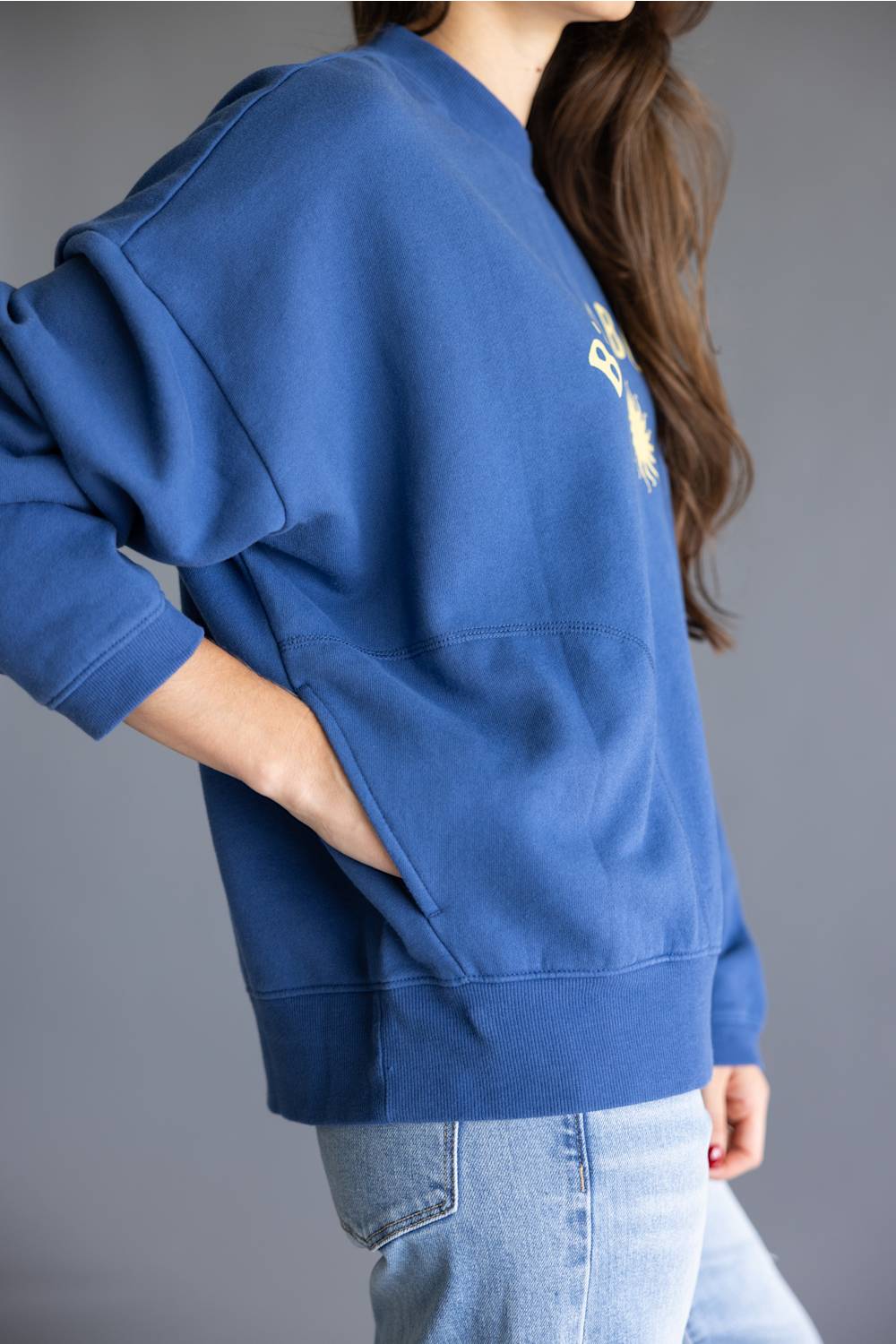 A person wearing an oversized blue sweatshirt with a small white graphic on the chest, paired with light blue jeans, shown against a plain gray studio background.