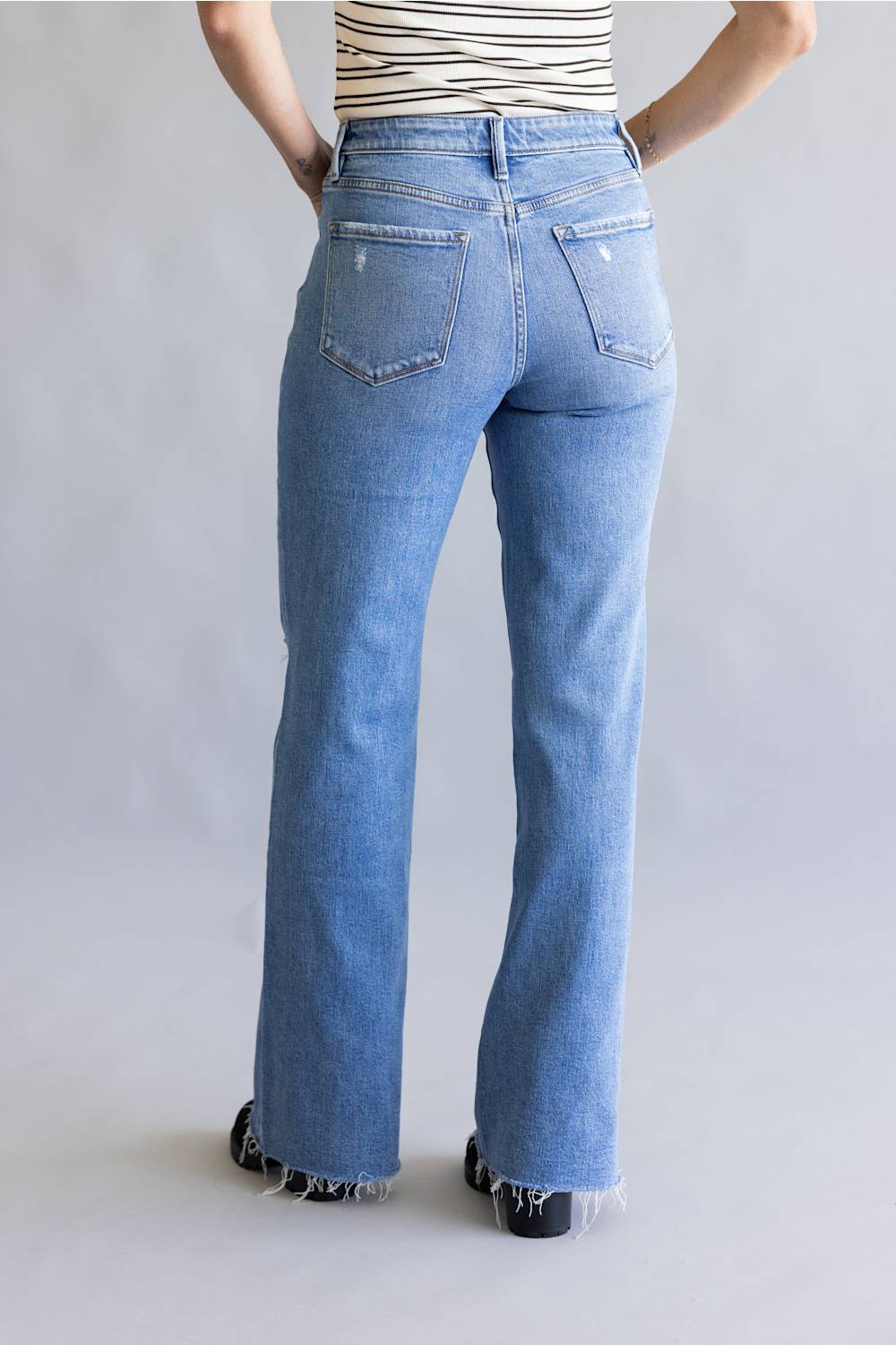A person shown from the back wearing light blue high-waisted flare jeans with frayed hems, paired with a striped top and black shoes, against a light gray studio background.
