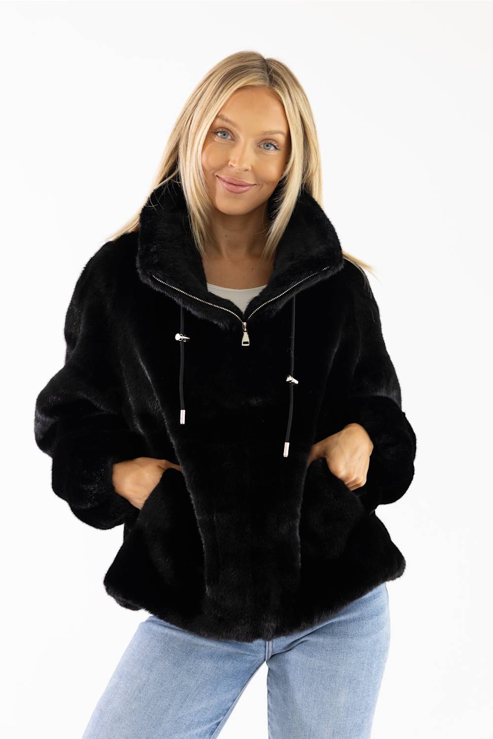 A woman wearing a black faux fur hooded jacket with a drawstring and zipper, paired with light blue jeans, against a plain white background.